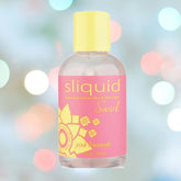 A bottle of Pink Lemonade Flavored Lubricant by Sliquid, a natural intimate lubricant, is placed against a softly blurred background with colorful bokeh lights. The 4.2oz water-based lubricant features a yellow cap and decorative patterns in yellow and pink on its label. It’s glycerin and paraben-free for your comfort.